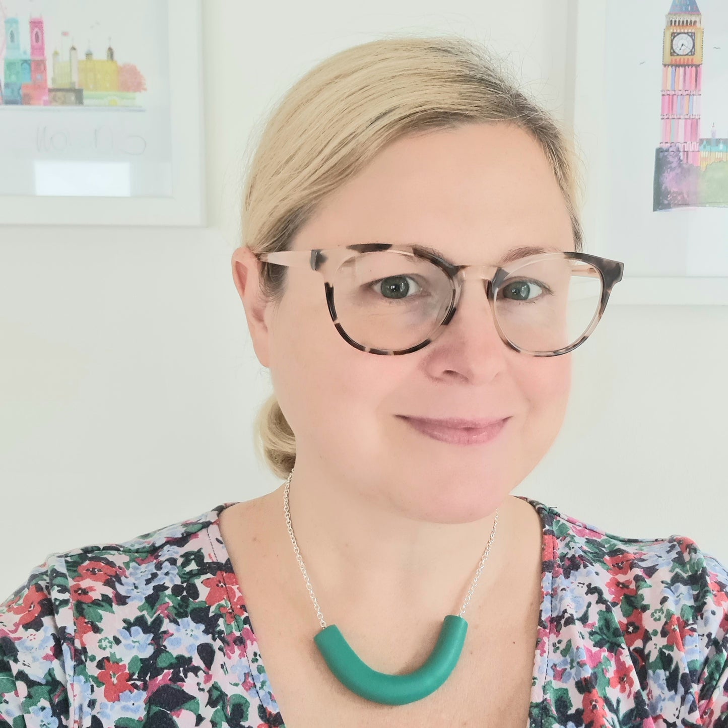 Amy Ayton is wearing a chunky green necklace, a blue & green floral dress and brown glasses.