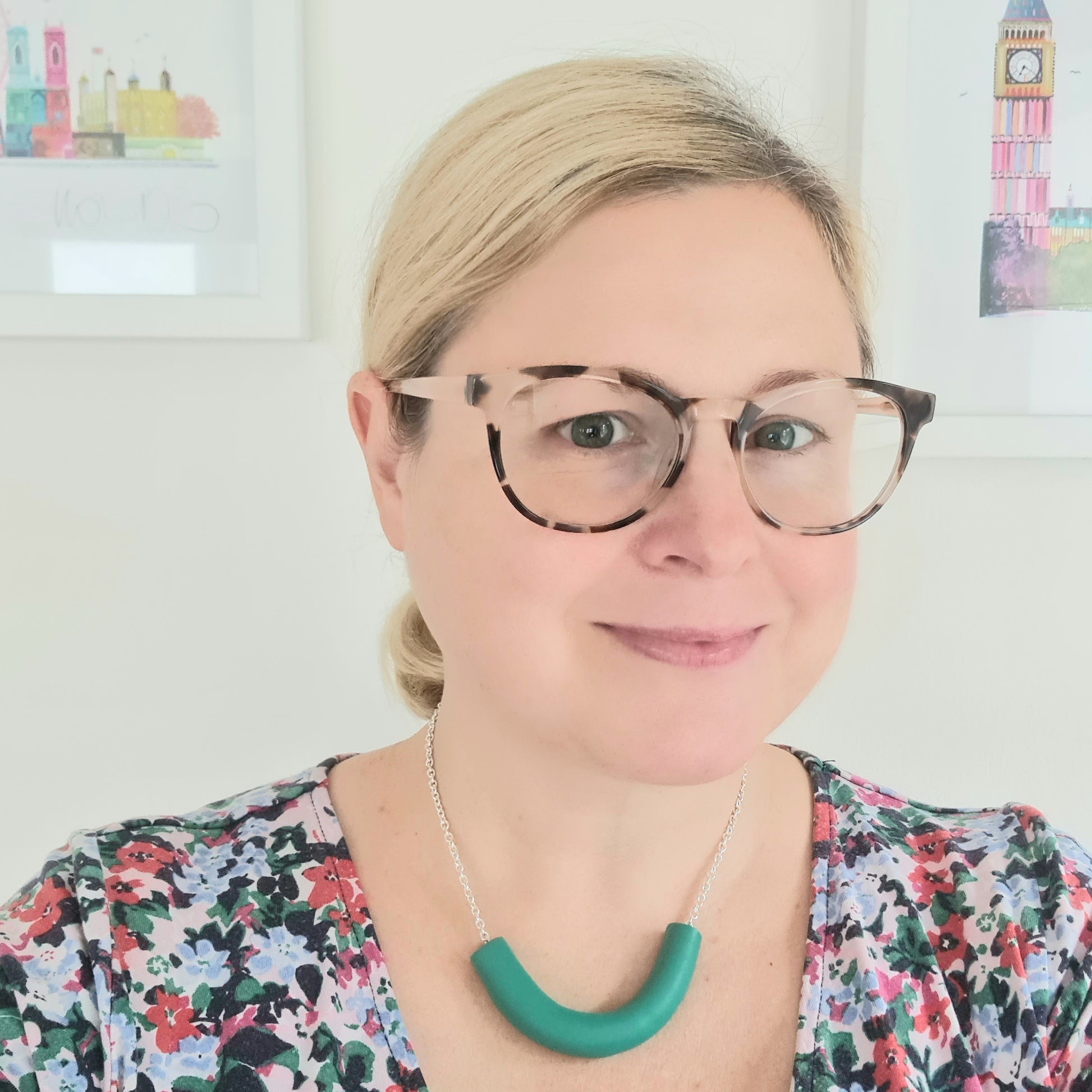 Amy Ayton is wearing a chunky green necklace, a blue & green floral dress and brown glasses.