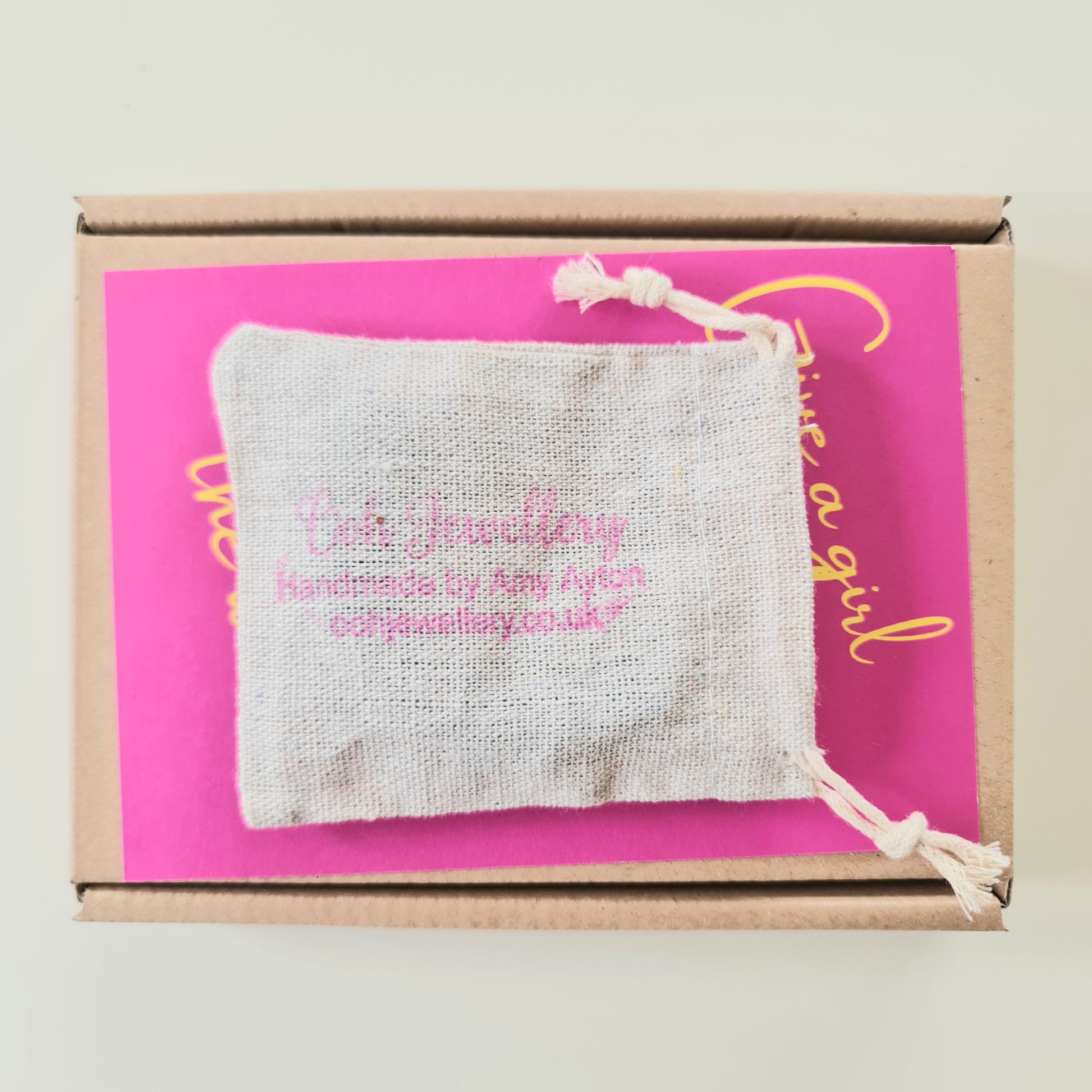 A brown postal box with a cream coloured hessian jewellery pouch on top of it. The pouch has 'Ooh Jewellery- handmade by Amy Ayton' stamped on the front in pink ink.