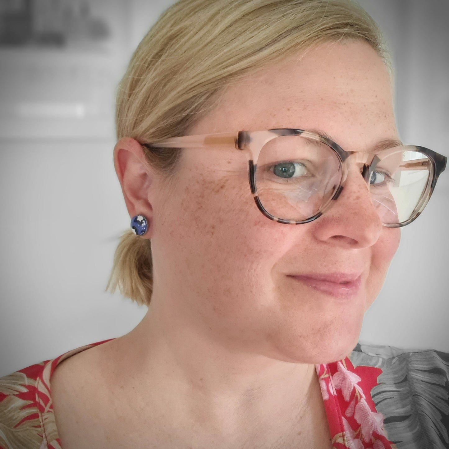 A picture of Amy Ayton wearing a pair of handmade blue leopard print stud earrings. Amy is a white woman with medium length blonde hair tied back in a pony tail. She is wearing brown tortoiseshell glasses.