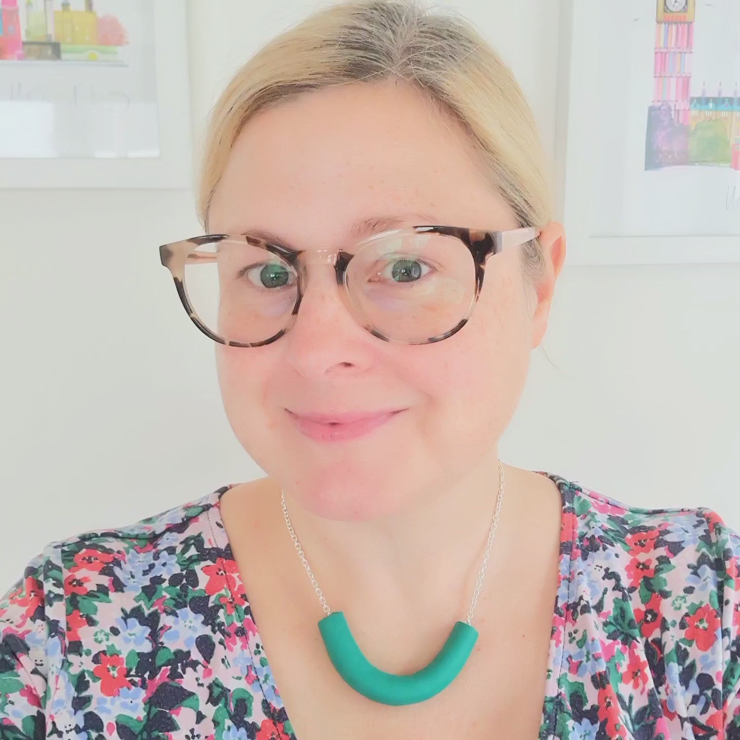 A video of Amy wearing a chunky green necklace. Amy is a white female with blonde hair tied back in a pony tail. She is wearing a blue and green floral dress and brown glasses.